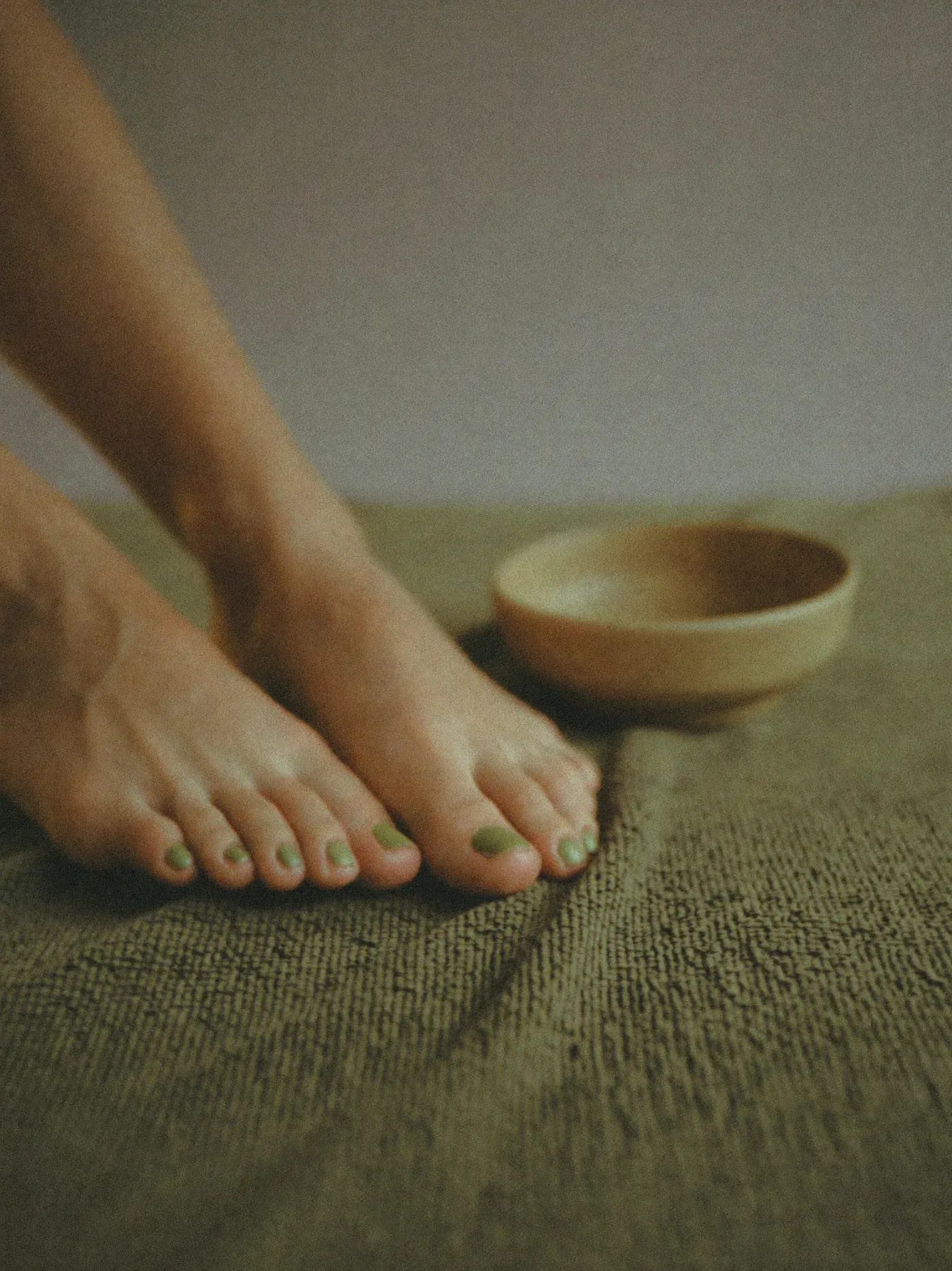 Feet with a sage-green pedicure on woven linen beside an empty ceramic bowl